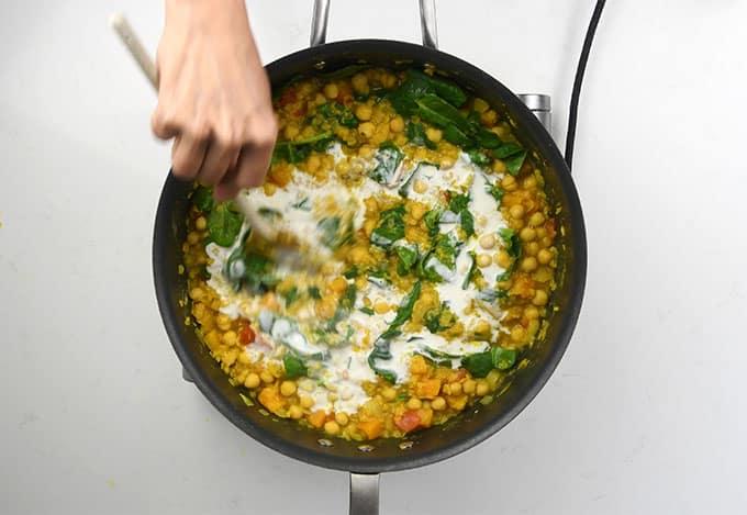 A hand stirs a vegan mixture of chickpeas, spinach, and creamy sauce in a black pan on a white surface, reminiscent of an Indian red lentil dal.