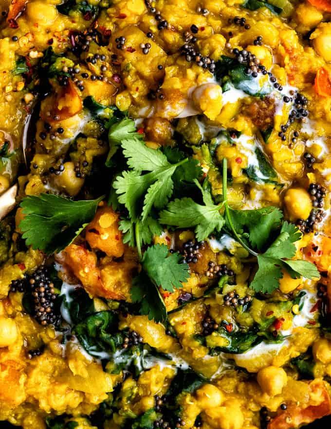 A close-up of hearty, creamy yellow and red lentil dal with chickpeas, spinach, black mustard seeds, and fresh cilantro garnish. Swirls of yogurt are visible on top, adding a touch of white to this vibrant and healthy dish.