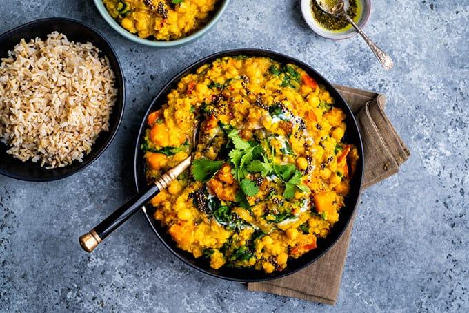 A bowl of vegan yellow lentil curry, reminiscent of classic Indian red lentil dal, garnished with fresh cilantro and black mustard seeds, served with brown rice and a small bowl of green chutney on the side.