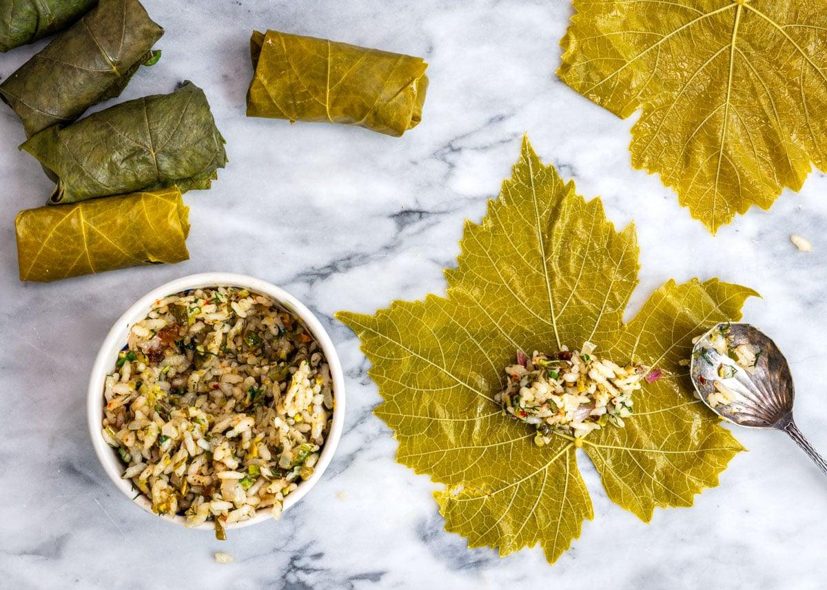 A marble surface with fresh grape leaves, some wrapped around rice filling, others unwrapped. A bowl of rice mixture and a spoon rest beside an unwrapped leaf with rice filling in the center, ready to be rolled.