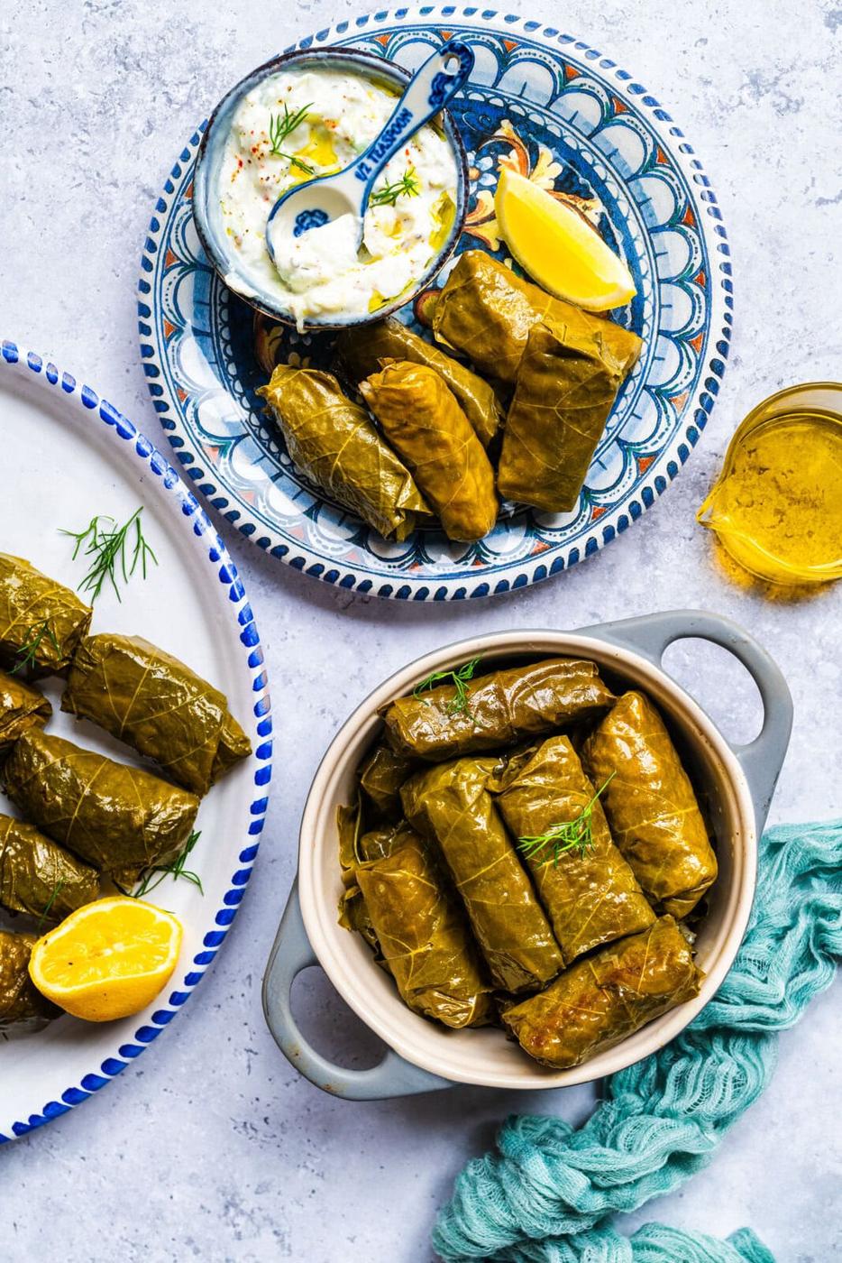 Plates and a bowl filled with stuffed grape leaves (dolmas), served with lemon wedges, a small dish of creamy dip topped with herbs, and a small pitcher of olive oil on a light textured surface.