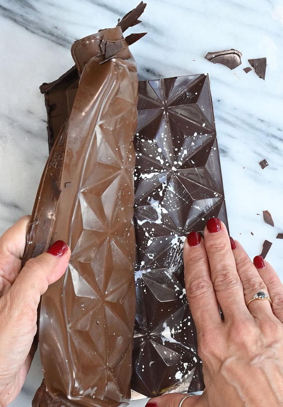Two hands with red nail polish unwrap a bar of dark chocolate on a marble surface, revealing geometric patterns on the chocolate and some white powder dusted on top.