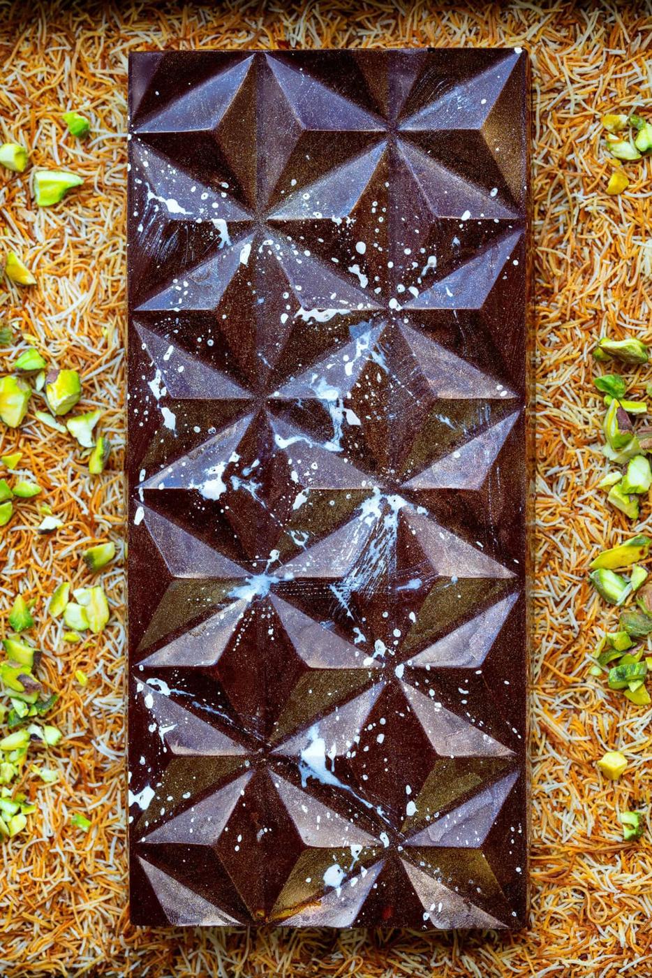 A glossy Dubai chocolate bar with a geometric pattern and white splatters rests on a bed of golden kataifi pastry and chopped green pistachios.