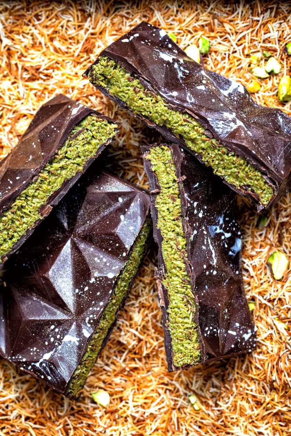 Three chocolate bars with geometric patterns are sliced to reveal a vibrant green pistachio filling, resting on a bed of golden shredded pastry and sprinkled with a few pistachio pieces.