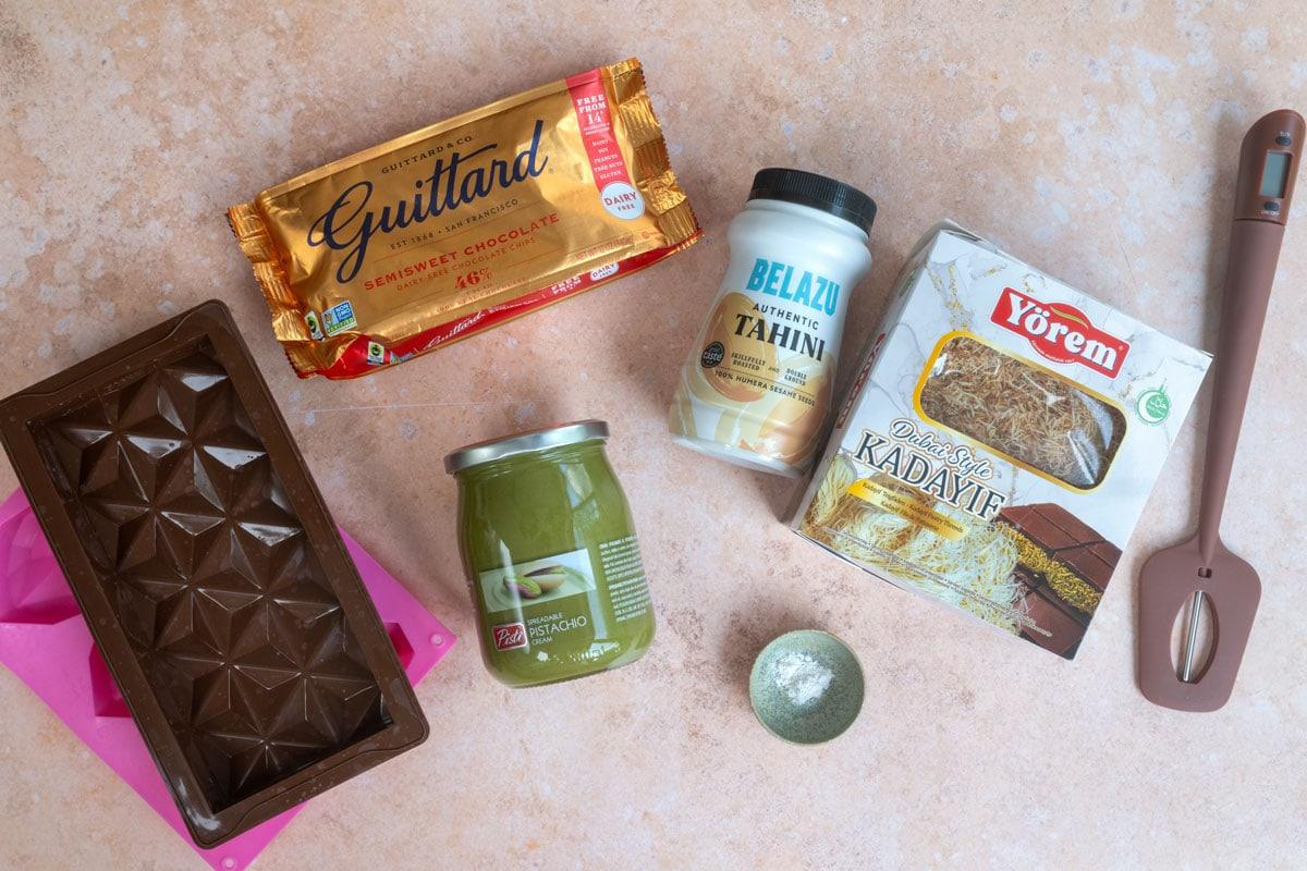 A selection of baking ingredients on a pink stone surface: a Guittard chocolate bar, Belazu tahini, pistachio spread, a box of kadayif pastry, a jar of matcha, a silicone spatula, and a geometric chocolate mold.