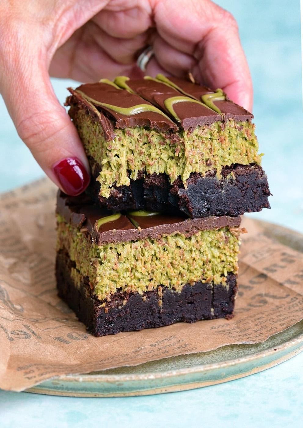A hand with red nail polish holds two stacked Dubai Chocolate brownies, featuring a dark chocolate base, green pistachio layer, and chocolate topping, placed on a plate with parchment paper.