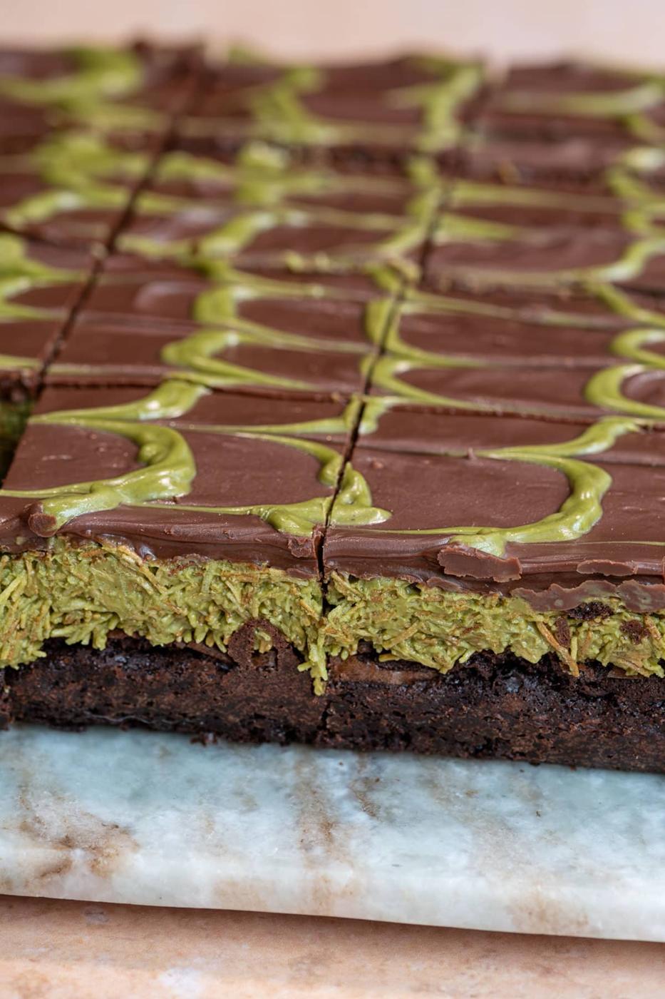 Close-up of sliced Dubai chocolate brownies with a thick pistachio kataifi layer and chocolate frosting, decorated with green swirls on top, arranged on a marble surface.