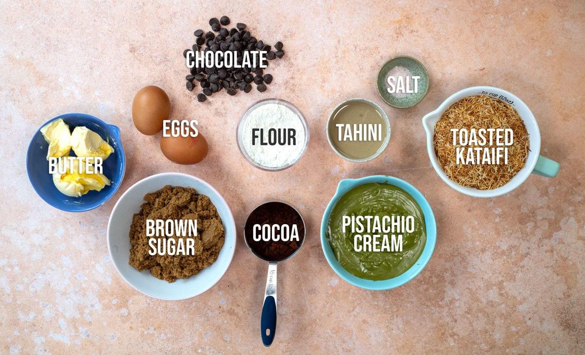 A top-down view of baking ingredients in bowls: chocolate chips, eggs, flour, tahini, salt, toasted kataifi, butter, brown sugar, cocoa powder, and pistachio cream, each labeled with text.