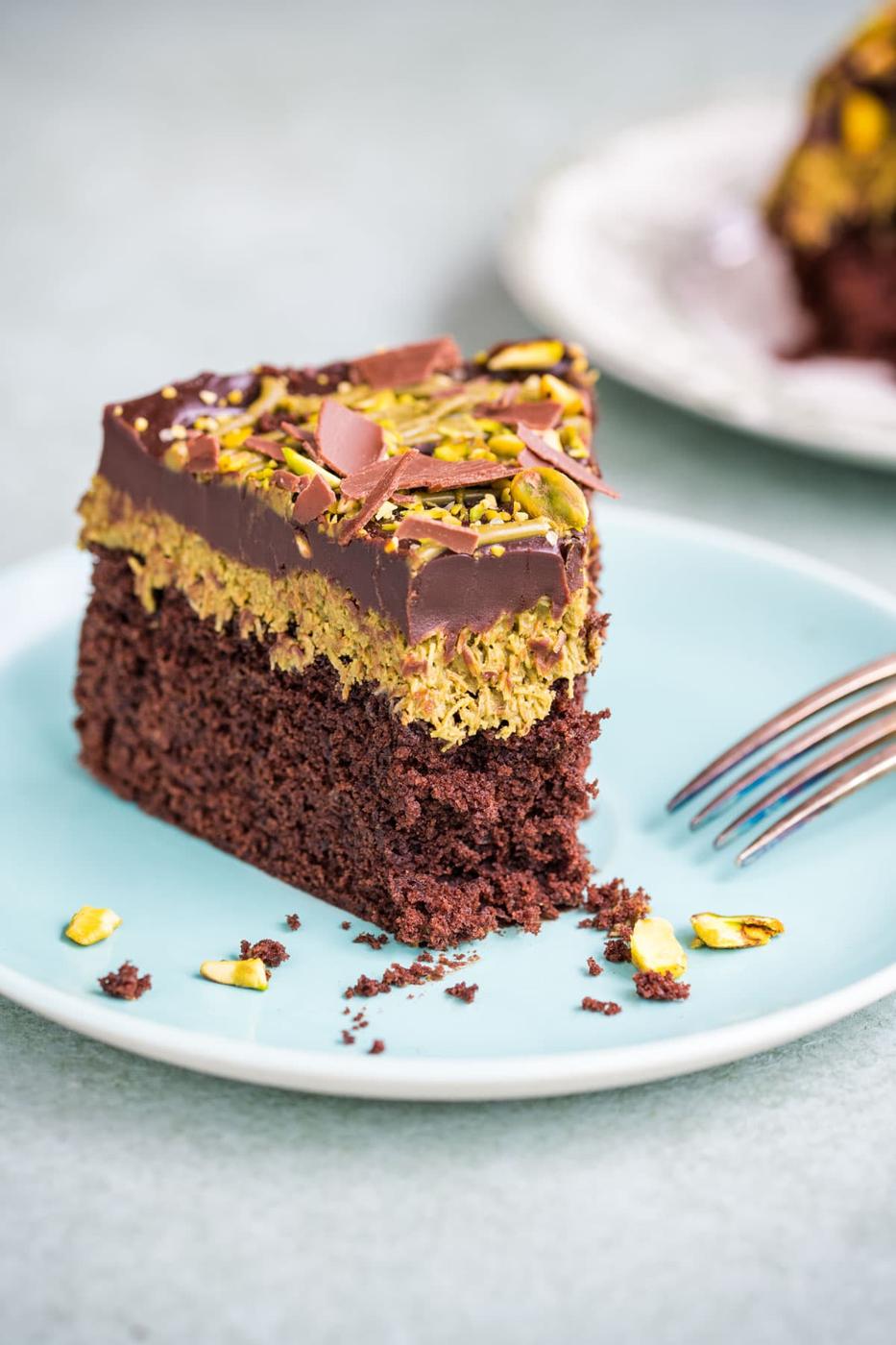 A slice of chocolate cake with a chocolate ganache and pistachio topping, served on a light blue plate with a fork and scattered pistachio pieces.