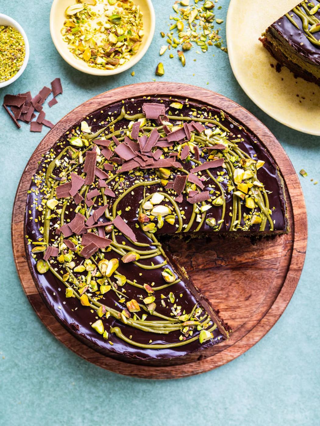 A round Dubai chocolate cake with glossy icing, topped with chopped pistachios and chocolate shavings, sits on a wooden board with a large slice missing. Bowls of pistachios are nearby on a light green surface.