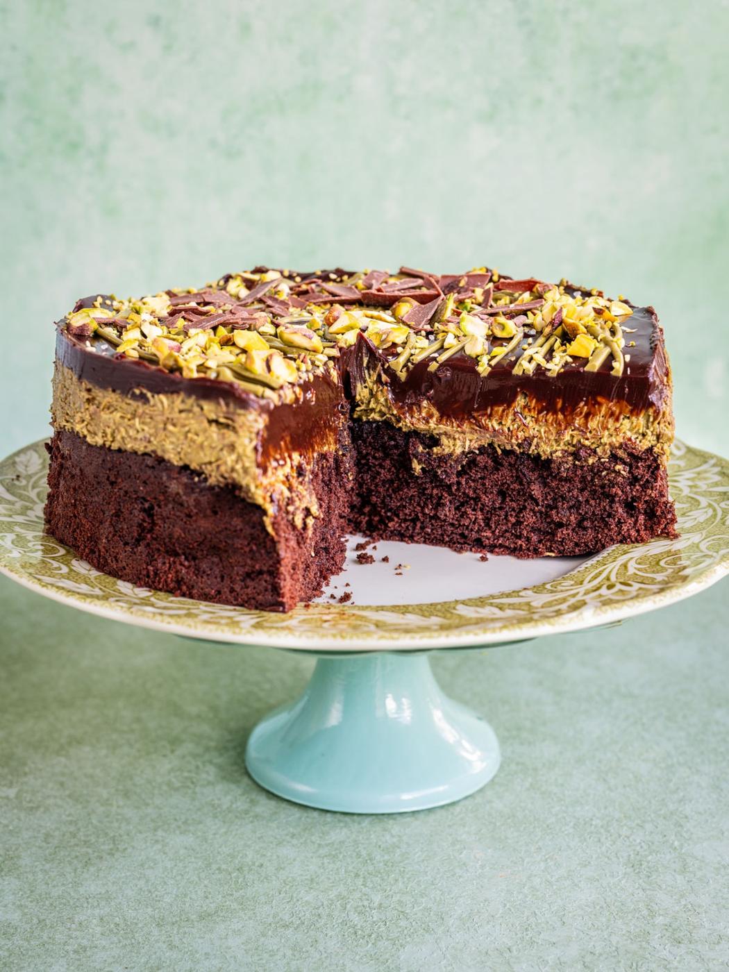 Dubai chocolate cake with a slice removed, topped with a thick chocolate layer, creamy frosting, and chopped nuts, displayed on a decorative cake stand against a light green background.