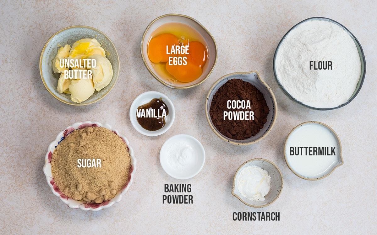 Top-down view of bowls containing ingredients for baking: unsalted butter, large eggs, flour, sugar, vanilla, cocoa powder, buttermilk, baking powder, and cornstarch, each labeled with white text.