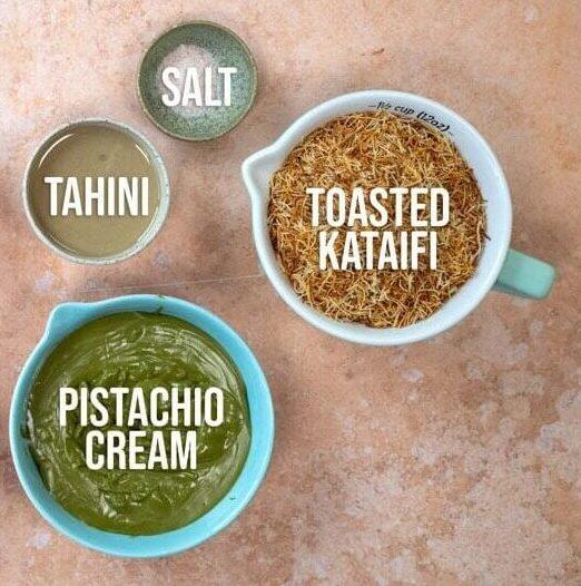 A top-down view of baking ingredients in bowls: chocolate chips, eggs, flour, tahini, salt, toasted kataifi, butter, brown sugar, cocoa powder, and pistachio cream, each labeled with text.