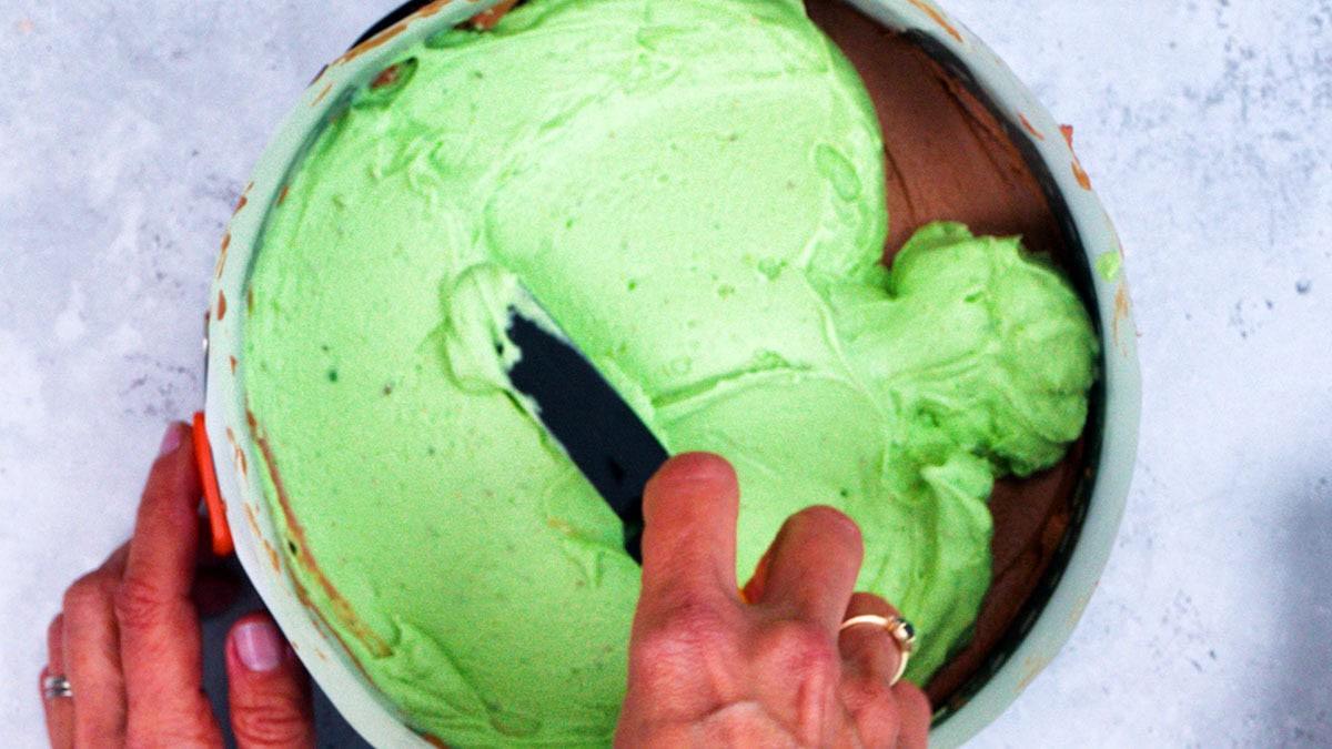 A person spreads light green pistachio cheesecake filling over a chocolate base in a round pan, using a spatula. The background is a light, textured surface.