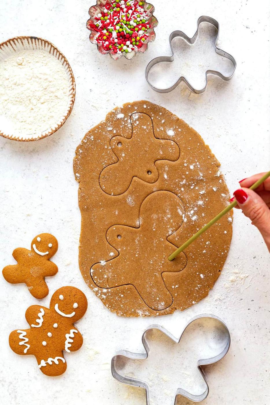 A hand uses a straw to make a hole in gingerbread cookies shaped like people. Cookie cutters, decorated gingerbread cookies, sprinkles, and flour are nearby on a white surface.