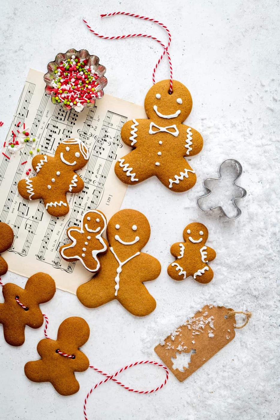 Easy homemade Gingerbread cookies, some decorated with white icing and red string loops, are arranged on a light surface with a music sheet, metal cookie cutter, sprinkles, and a blank brown gift tag nearby.