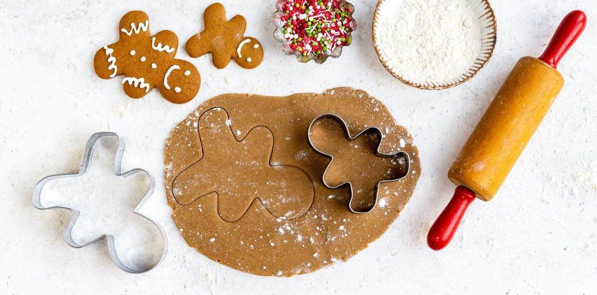 A gingerbread man cookie cutter sits on rolled-out dough, surrounded by festive Gingerbread cookies, colorful sprinkles, a bowl of flour, and a rolling pin—perfect for trying your favorite gingerbread cookies recipe on a red-striped cloth.
