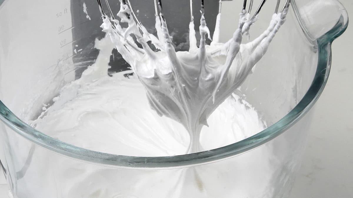 A glass mixing bowl with fluffy, white royal icing. A metal whisk attachment, lifted from the bowl, shows stiff peaks of the royal icing clinging to it.