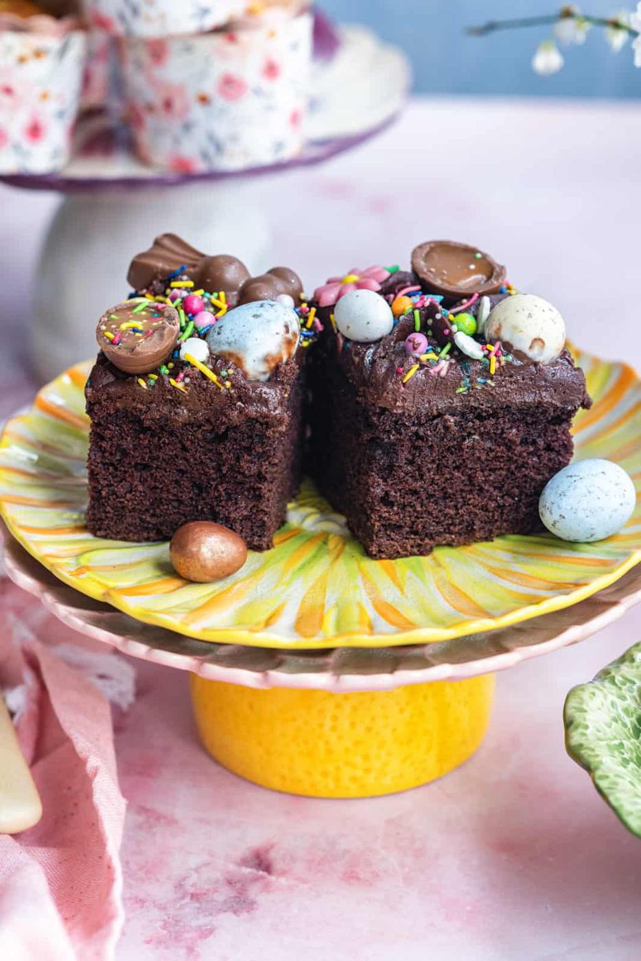 Two squares of Easter chocolate tray bake cake on a cake stand, easter egg candy decorations on top of the cake and on the side