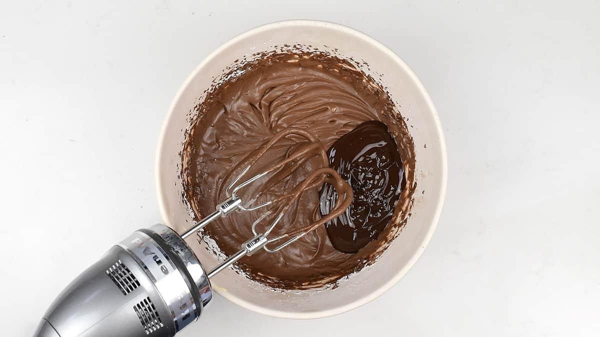 A bowl of chocolate frosting for a decadent traybake is being mixed with an electric hand mixer. The creamy, swirled mixture shows the mixer blades partially submerged against a pristine white background.