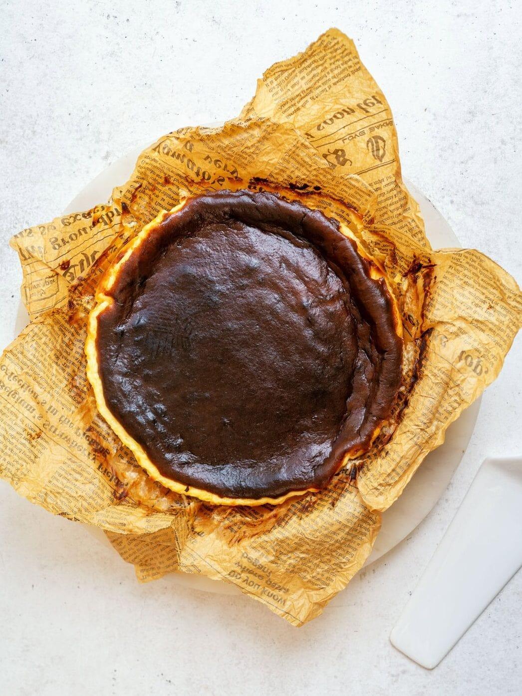 A burnt Basque cheesecake sits on a plate, lined with crinkled brown parchment paper printed with text, next to a white cake server on a light surface.
