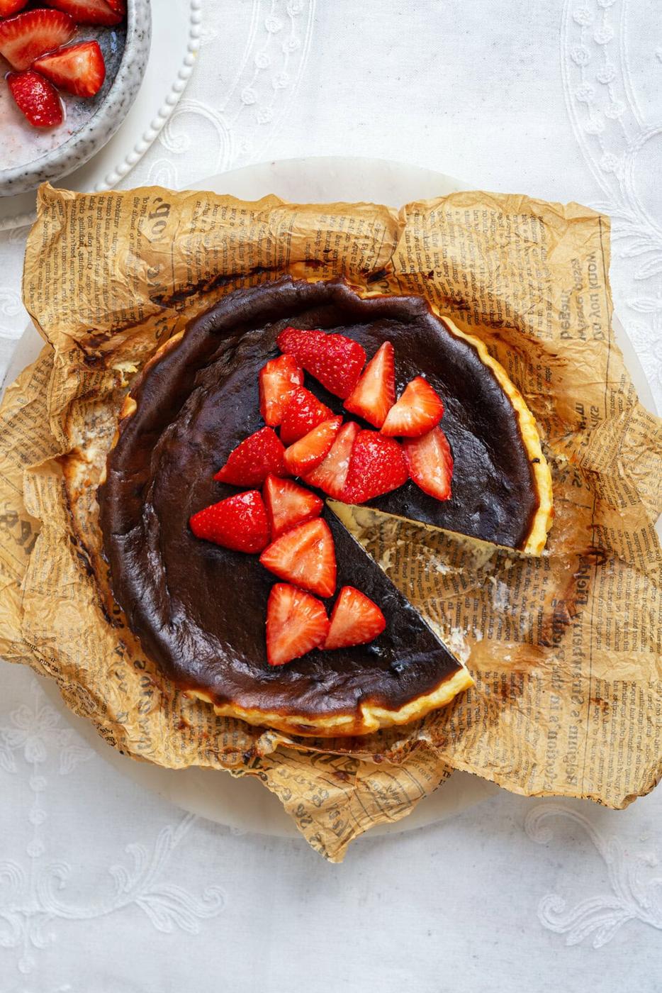 A rustic burnt cheesecake topped with fresh strawberry slices, set on brown parchment paper with a dark crust. A slice is missing, and a plate with more strawberries is visible in the corner.
