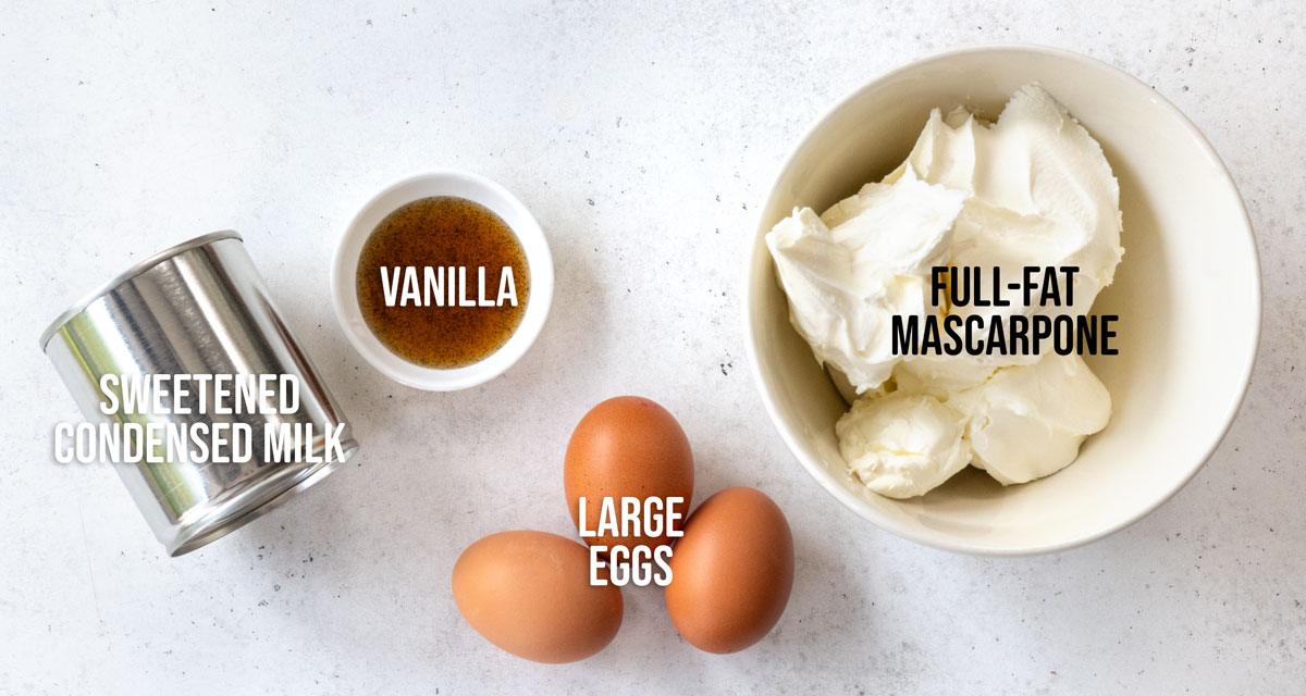 A can of sweetened condensed milk, a small bowl of vanilla, three large eggs, and a bowl of full-fat mascarpone cheese on a white surface, each labeled with text.