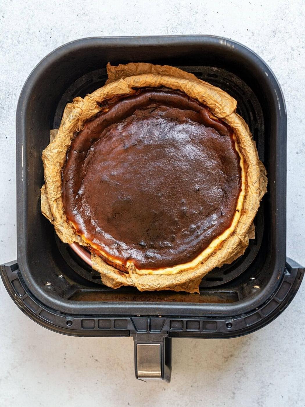 A dark, caramelized Basque cheesecake sits in an air fryer lined with brown parchment paper, viewed from above on a light-colored surface.