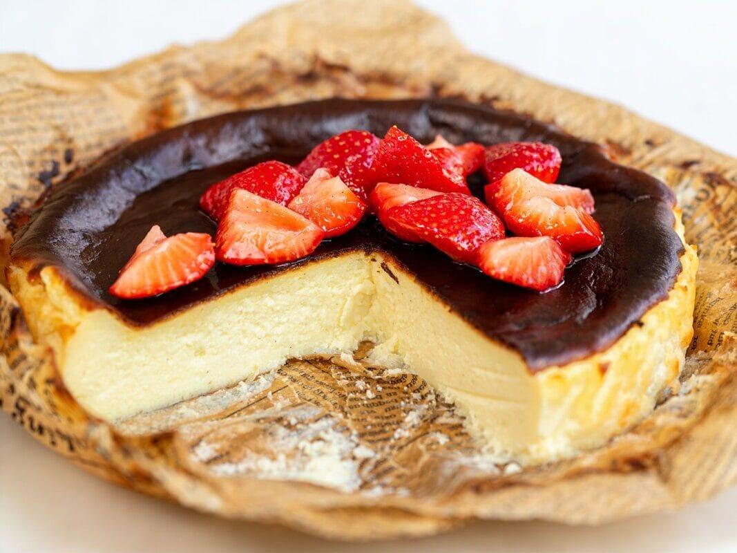A creamy cheesecake with a dark, caramelized top sits on parchment paper. A slice is missing, revealing the smooth interior. Fresh strawberry pieces are arranged on top as a garnish.