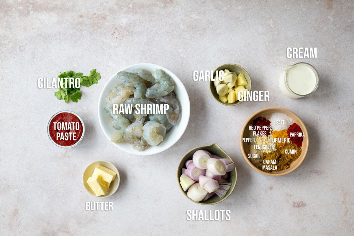 Various ingredients are arranged on a light surface, including a bowl of raw shrimp, cilantro, tomato paste, butter, shallots, garlic, ginger, cream, and a bowl of labeled spices like turmeric, cumin, and paprika.
