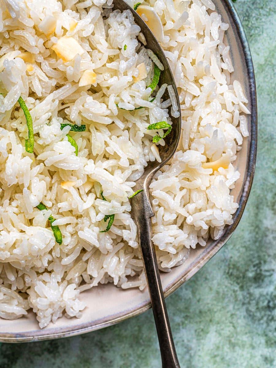 A close-up of a bowl of fluffy coconut rice garnished with thin green herbs, with a metal spoon resting inside the bowl. The background is a textured light green surface.