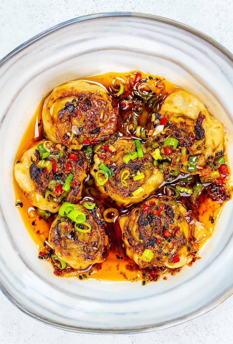 Six pan-fried dumplings topped with chopped green onions and red chilies sit in a white bowl, surrounded by a spicy chili oil sauce with visible pepper flakes and herbs.