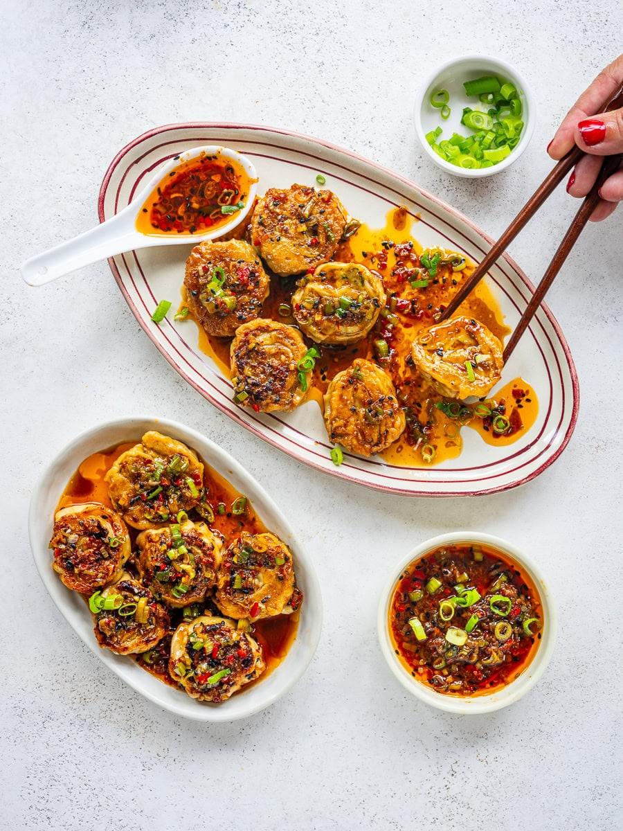 Two oval plates of stuffed eggplant rounds topped with chili oil and chopped green onions, with a bowl of dipping sauce and a small bowl of green onions nearby; a hand holds chopsticks picking up a piece.