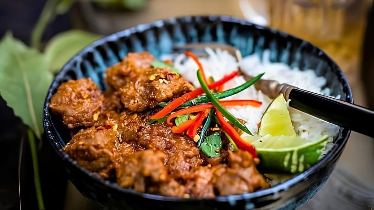 Lamb bhuna served over rice in a black bowl with sliced chilies, lime wedges and fresh herbs, with a fork resting inside.