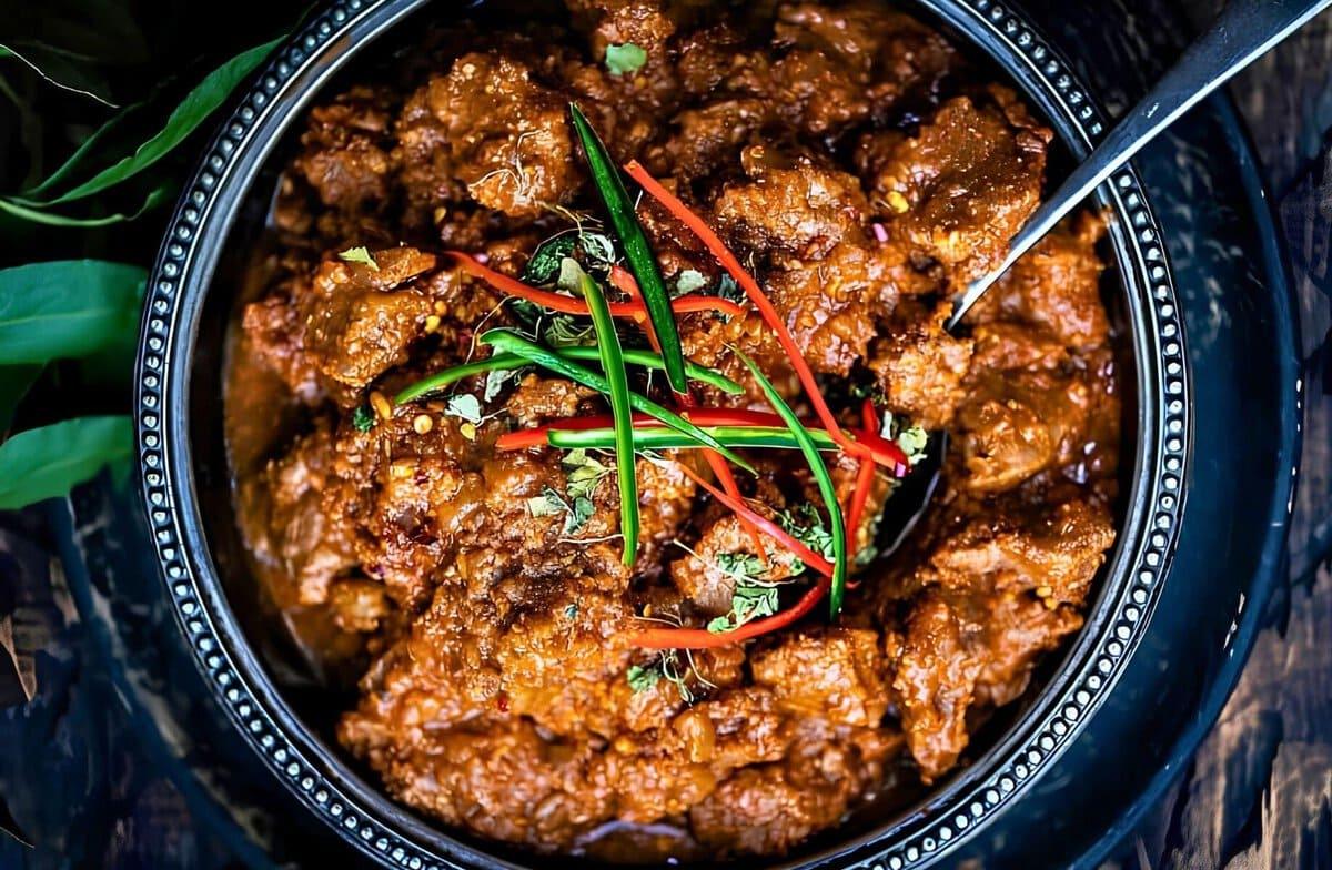 A black bowl of lamb bhuna garnished with thinly sliced red and green chilies and chopped herbs, with a spoon resting inside.
