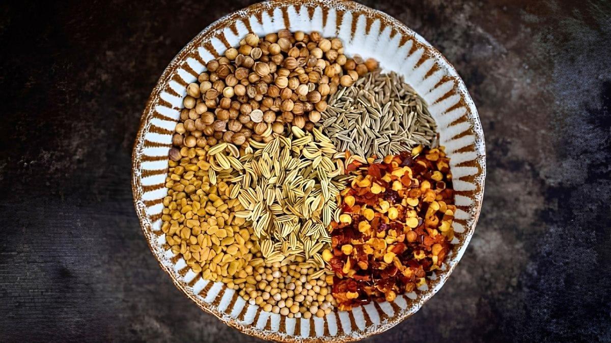A bowl of whole bhuna spices – coriander, cumin, fennel and mustard seeds with red chili flakes – arranged in separate sections.