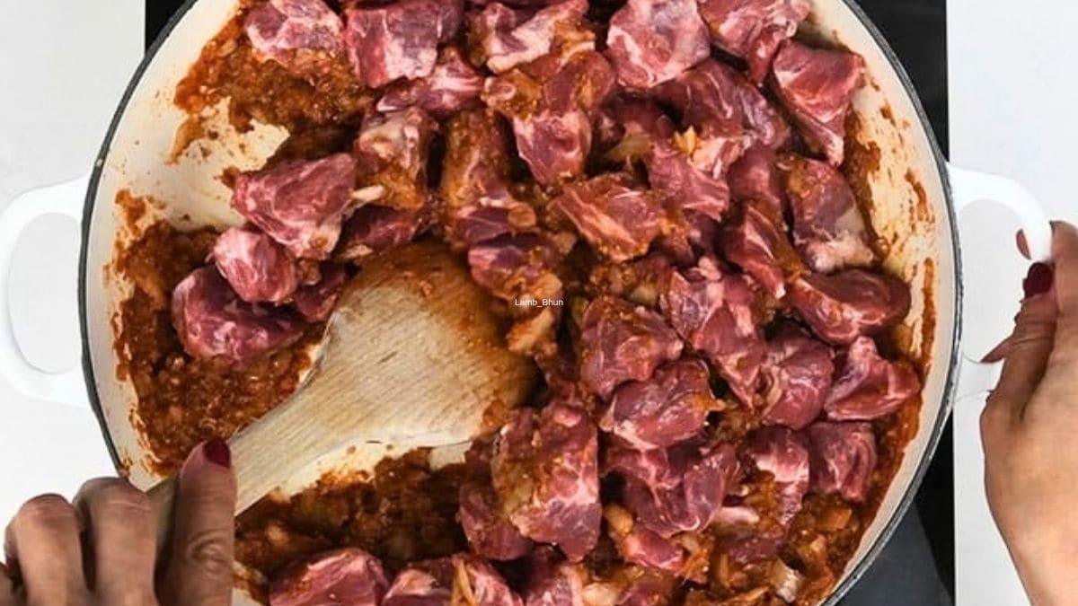 Stirring cubes of raw lamb into the spiced tomato masala in a white pot with a wooden spoon.
