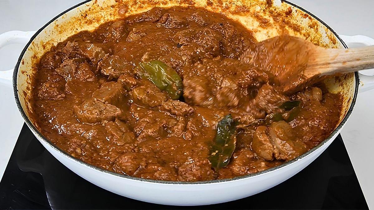 Lamb bhuna simmering in a white pot with bay leaves, the thick sauce clinging to the meat, being stirred with a wooden spoon.