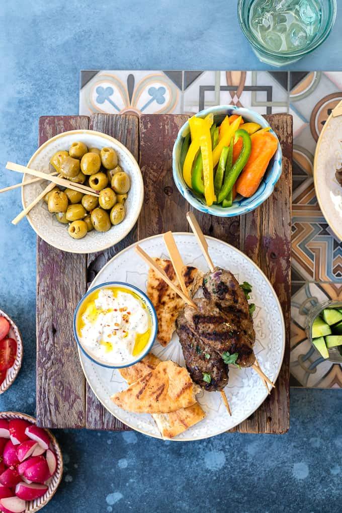 Lamb kofta on a plate with pitta bread and tzatziki surrounded by small plates of vegetables and olives