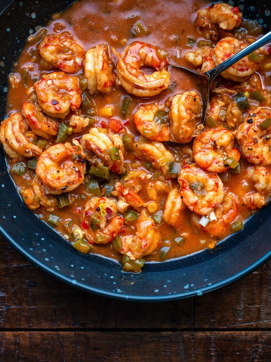 A close-up of a blue bowl filled with Cajun shrimp etouffee in a spicy, reddish-brown sauce with diced green peppers, and a spoon resting in the dish, all on a wooden surface.