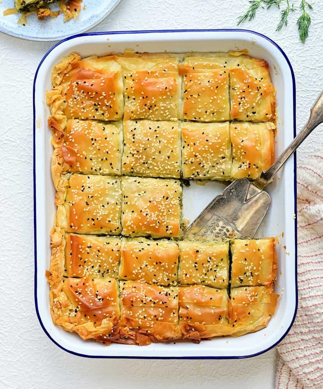 A rectangular baking dish with golden-brown, sesame-topped Greek spanakopita cut into squares. A serving spatula rests in the dish, with one spinach pie slice missing. Fresh dill and a plate with a serving are nearby.