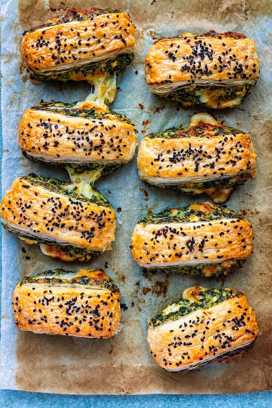 Eight golden Spinach Feta Puff Pastry Rolls filled with melted cheese and green pesto, topped with black sesame seeds, arranged on parchment paper. The flaky layers reveal some cheese deliciously oozing out.