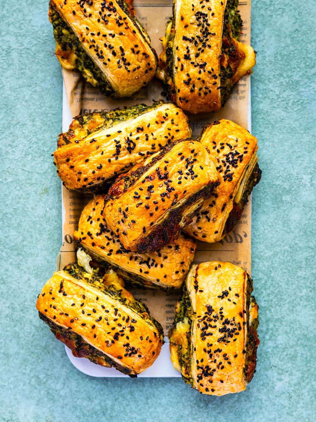 A platter of golden, flaky Spinach Feta Puff Pastry Rolls filled with green pesto and cheese, topped with black sesame seeds, displayed on parchment paper over a light blue surface.