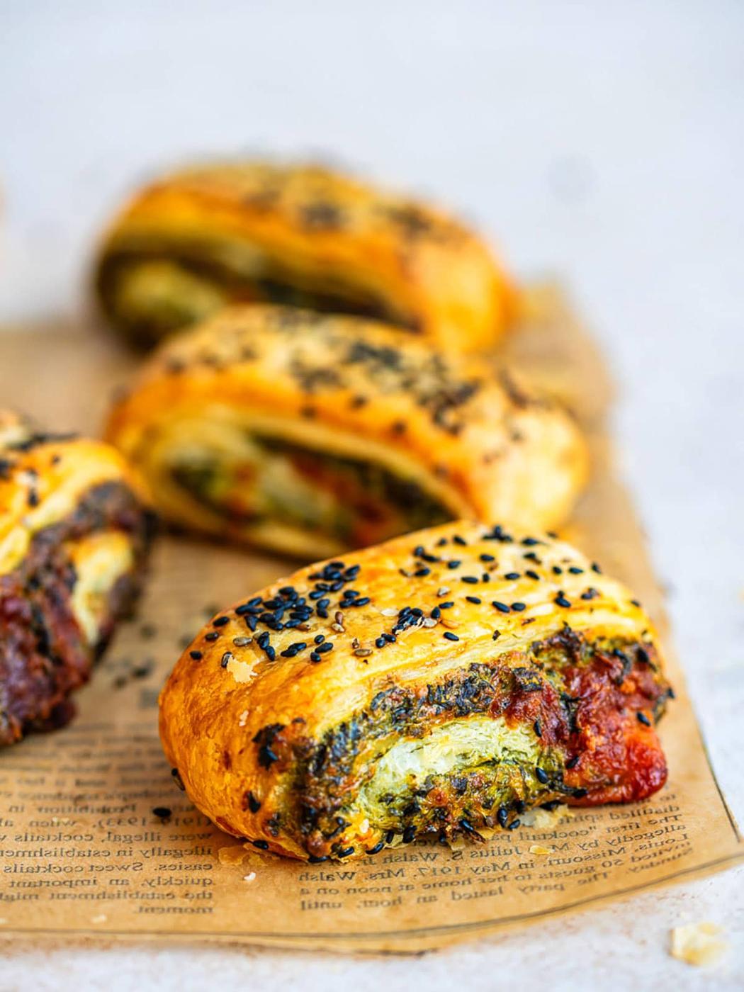 Golden Spinach Feta Puff Pastry Rolls filled with a green and red vegetable mixture, topped with black sesame seeds, rest on printed parchment paper. Three rolls are in the background, one in focus at the front.