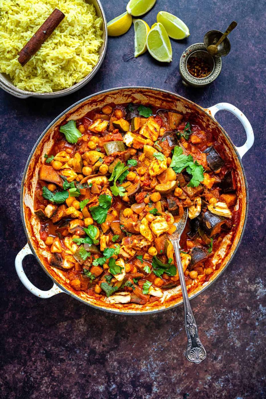 A pan filled with a vibrant vegetarian vegetable curry and bean stew, garnished with herbs, sits beside a bowl of yellow rice, lime wedges, and small dishes of spices on a dark surface. A spoon rests in the stew.