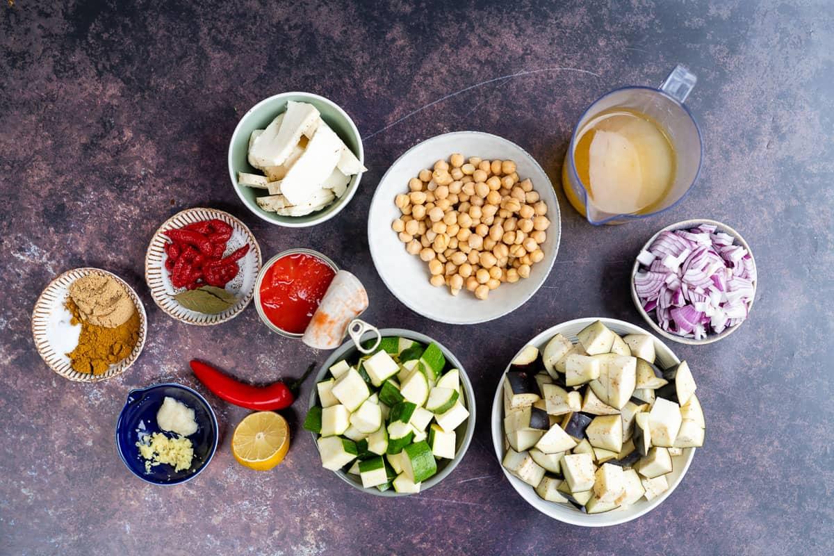 Assorted cooking ingredients for a vegetable curry in bowls on a dark surface, including chopped eggplant, zucchini, red onion, chickpeas, tofu, red chili, lemon, garlic, ginger, tomato, roasted red pepper, spices and a jug of broth. Perfect for a vegetarian dish.
