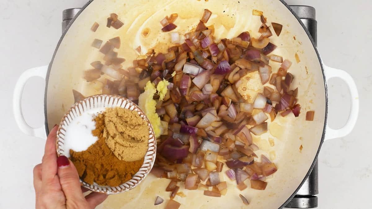 A hand holds a small bowl of ground spices, salt, and a yellow paste over a pot of sautéed chopped red onions on a stovetop, preparing the base for a flavorful vegetarian vegetable curry.