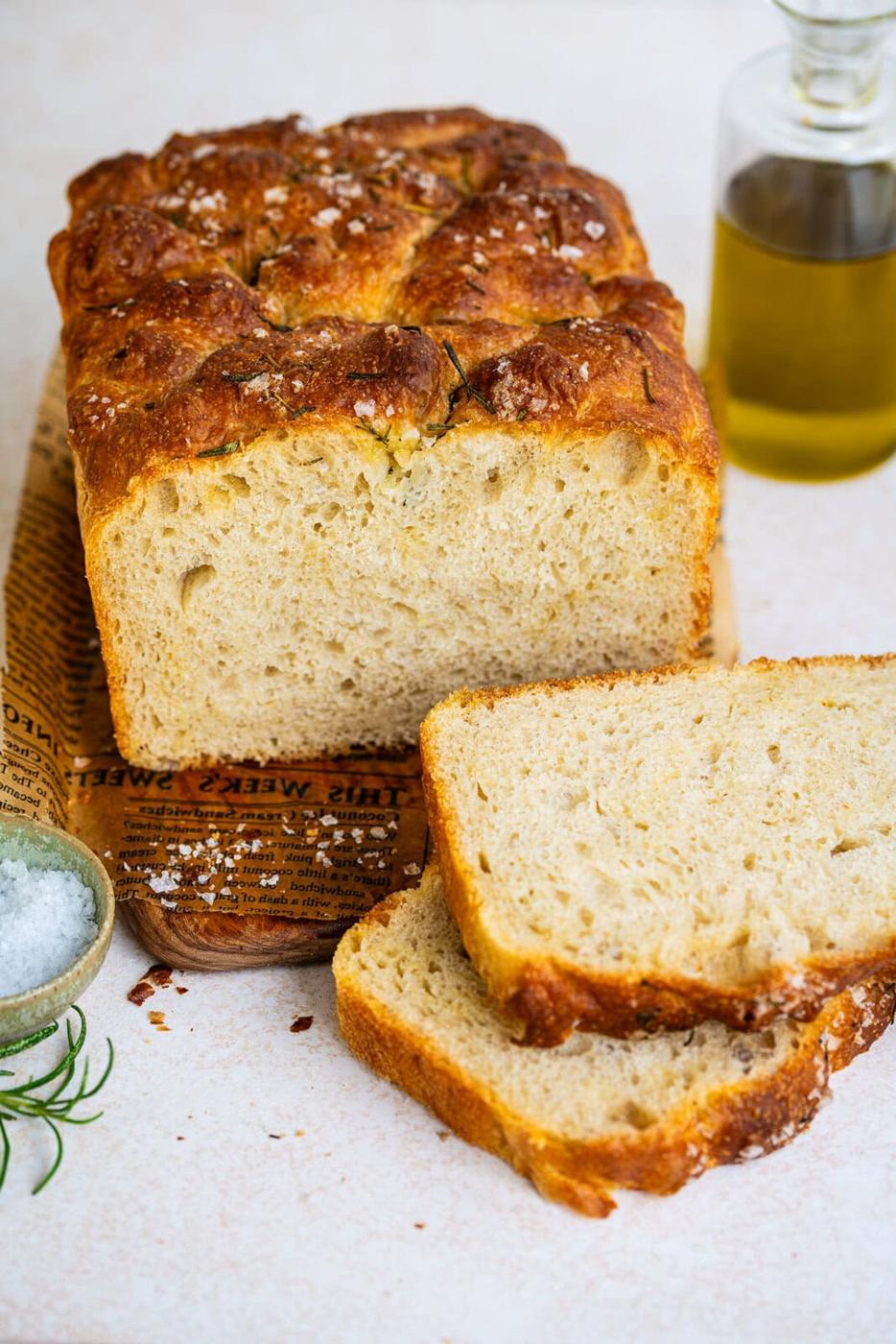 A loaf of freshly baked focaccia loaf bread with a golden crust sits on a wooden board, two slices cut in front. A small bowl of salt, a sprig of rosemary, and a bottle of olive oil are nearby.