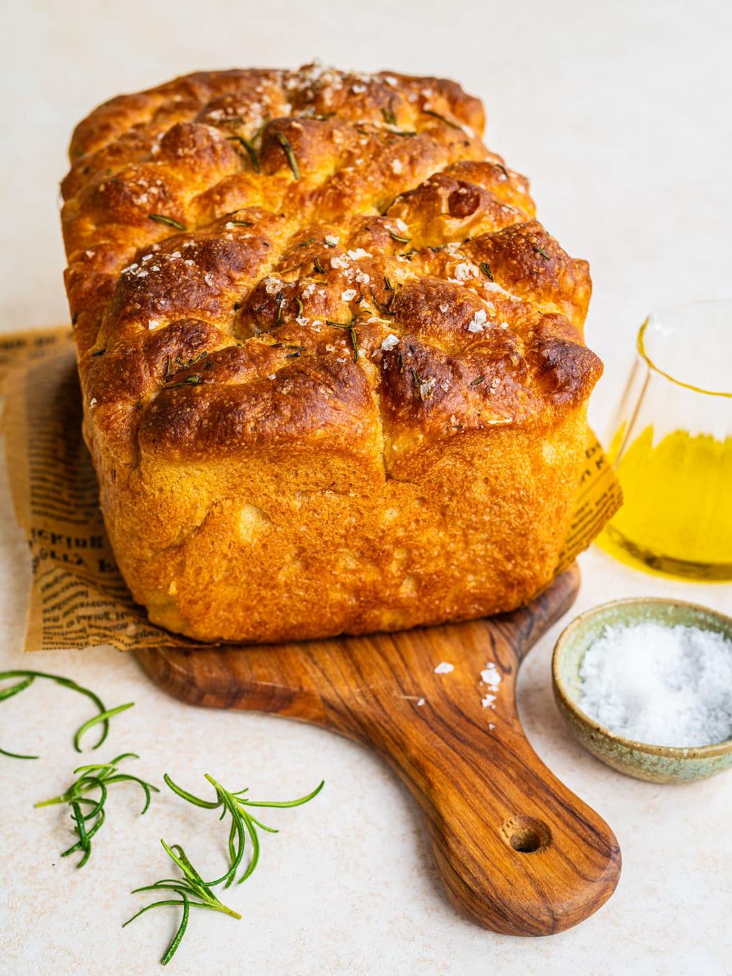 A golden, fluffy loaf of focaccia bread topped with sea salt and rosemary sits on a wooden board, with a bowl of salt, fresh rosemary sprigs, and a glass of olive oil nearby.