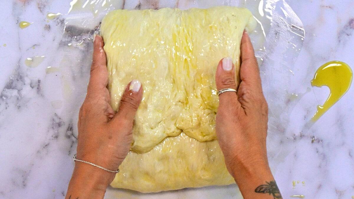 Hands with rings and a bracelet stretch oily dough on a marble surface, with some oil drizzled nearby and clear plastic wrap underneath.